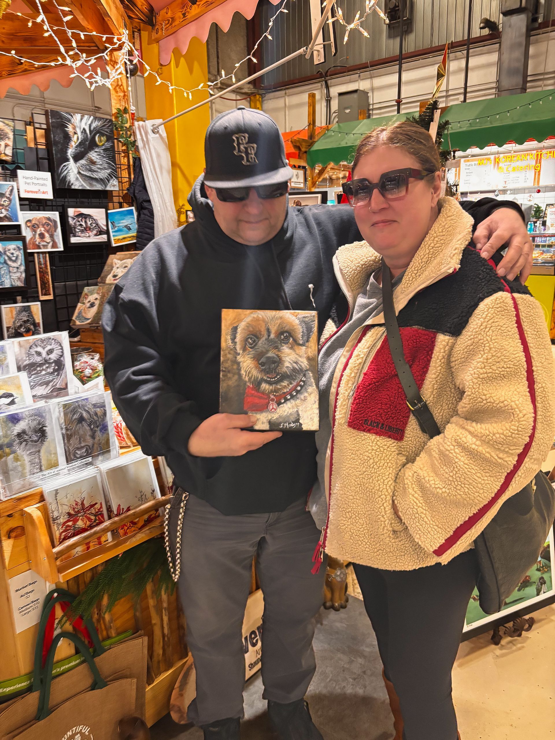 Man and woman pose holding dog portrait at a booth with art.