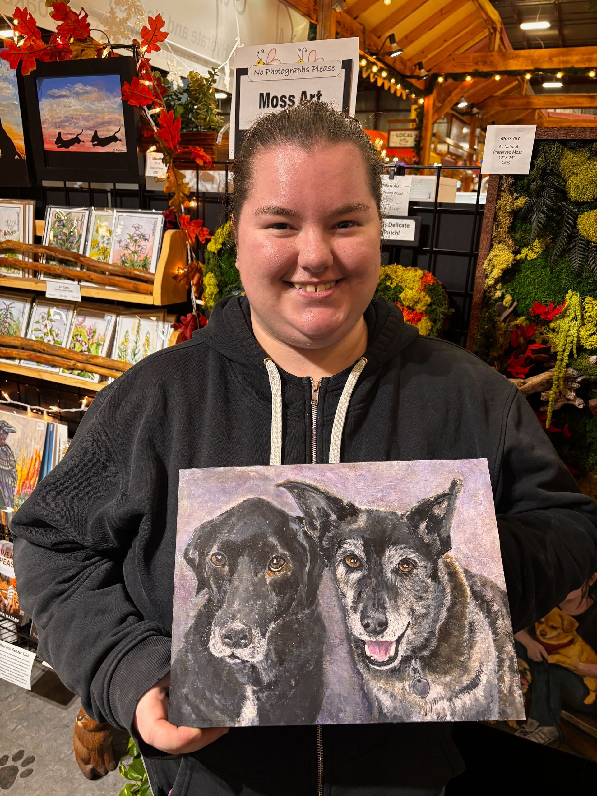 Woman smiling, holding a painting of two dogs. Art displayed in a shop setting.