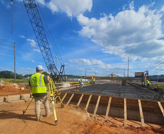 Surveyor in safety vest using a transit on a road construction site with a steamroller in the background.
