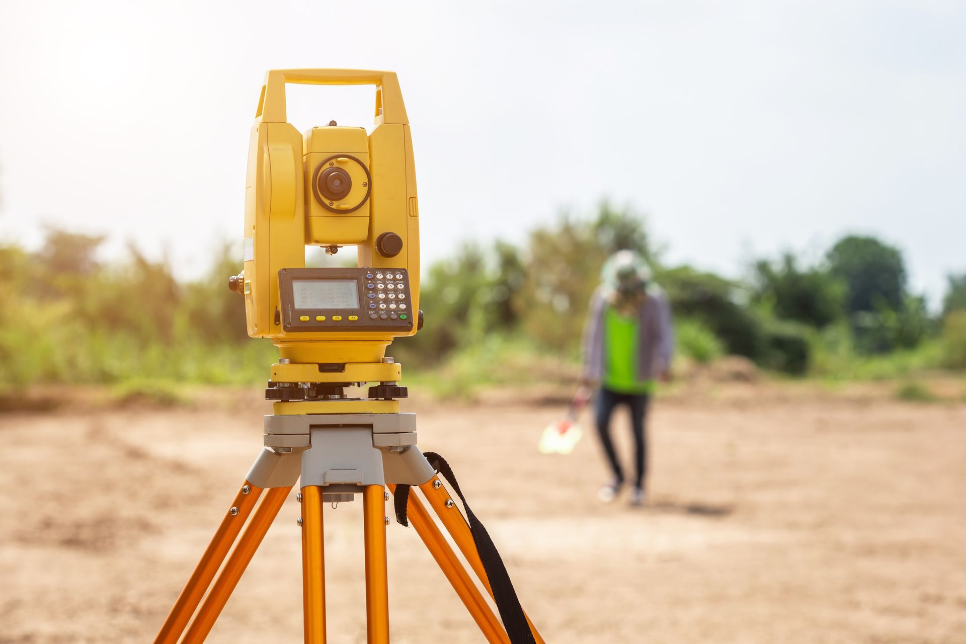 A yellow total station on a tripod in the foreground with a surveyor in a safety vest working onsite