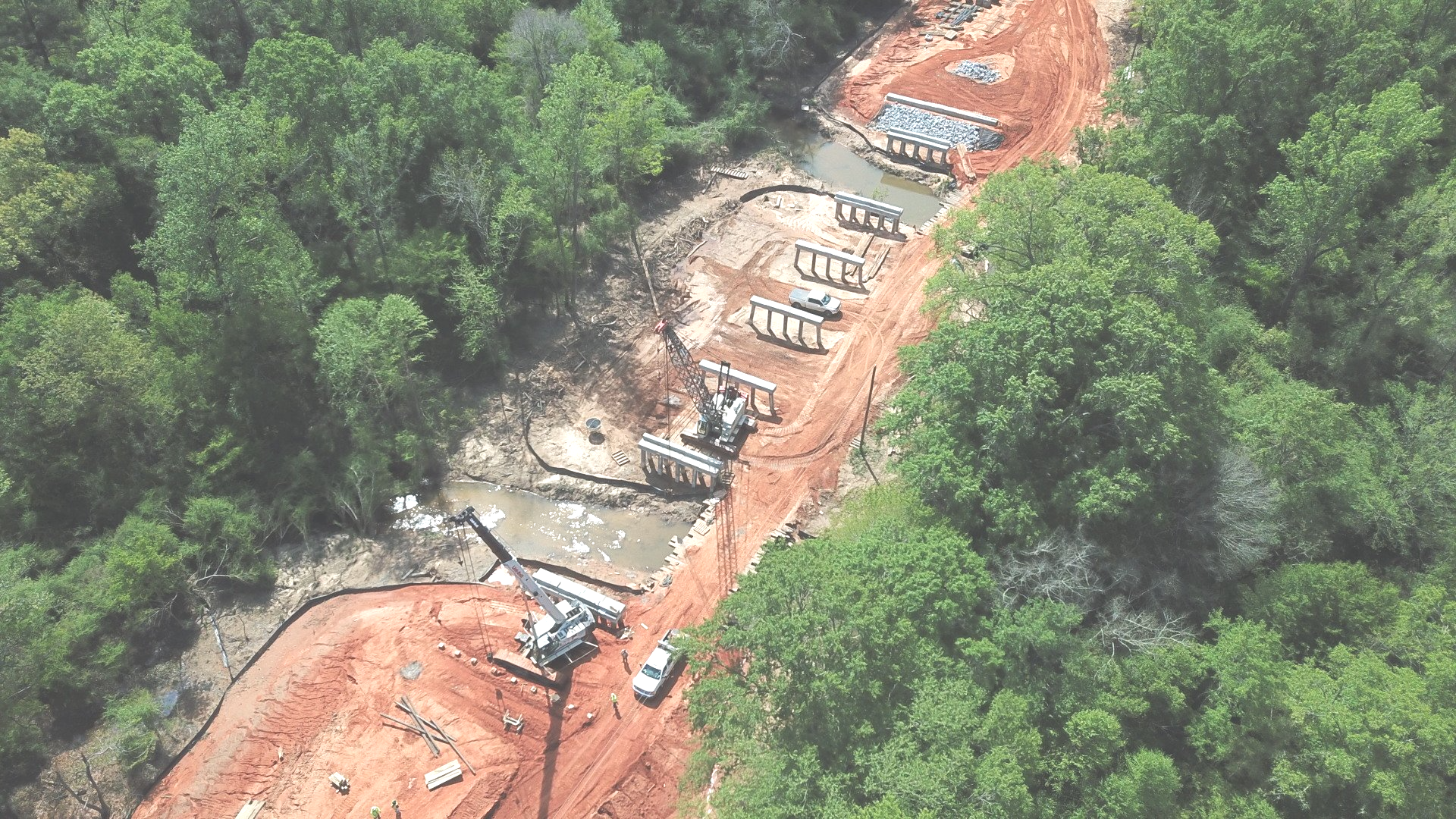 Aerial view of forest construction site with cranes and vehicles. Construction site in a forest clearing, with bridge supports in water and earthmoving equipment.