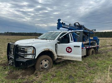 Two men operating a drilling rig mounted on a truck in a field. Cloudy sky, roadside setting.