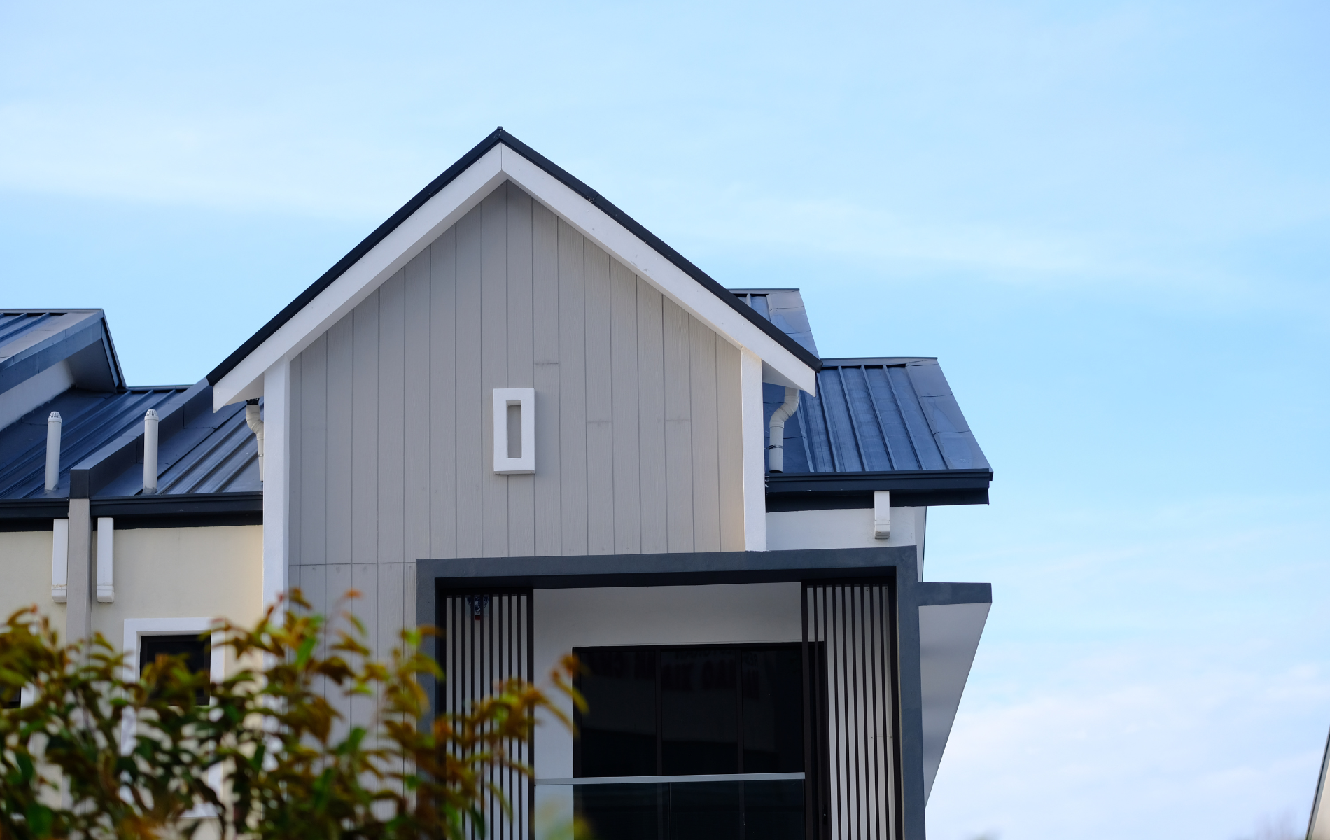 A modern house exterior featuring a grey vertical-paneled gable, a dark blue metal roof, and a shaded front entrance.