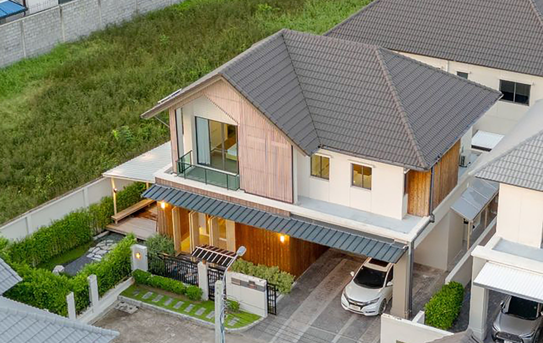 Aerial view of a two-story modern house with a dark tiled roof, wooden paneling, a carport, and a small garden path.
