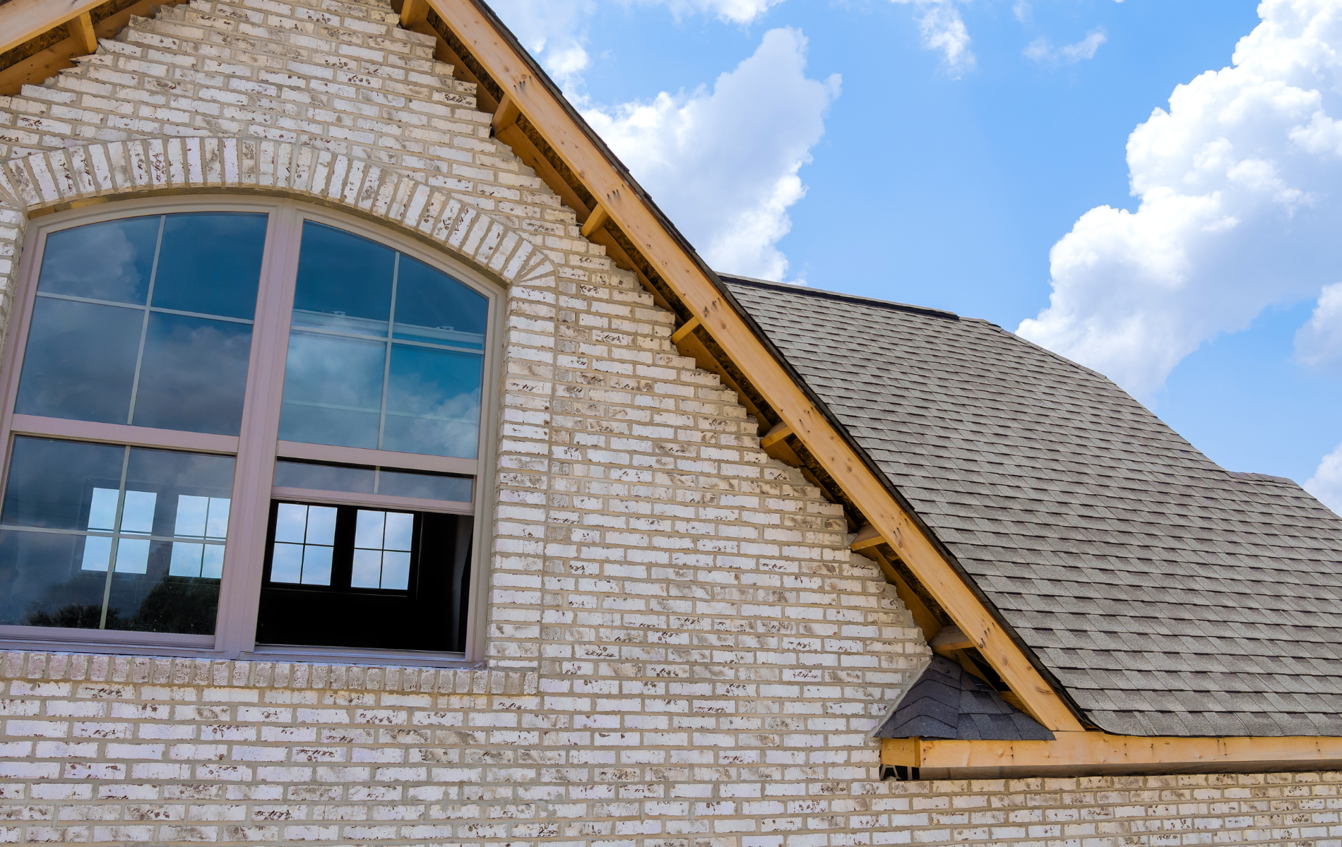 Close-up of a brick house under construction with a large arched window and a shingled roof against a blue sky.