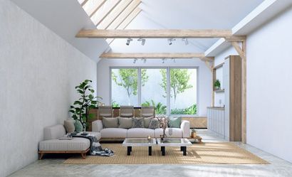 A sunlit living room with white walls, a large sectional sofa, wooden ceiling beams, and a view of greenery through windows.