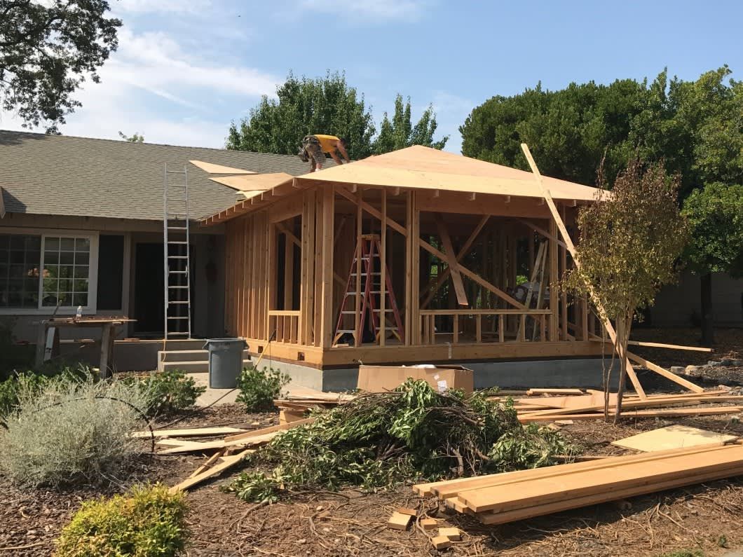 A wooden house addition under construction with a frame, plywood roof, and ladders, adjacent to an existing house.