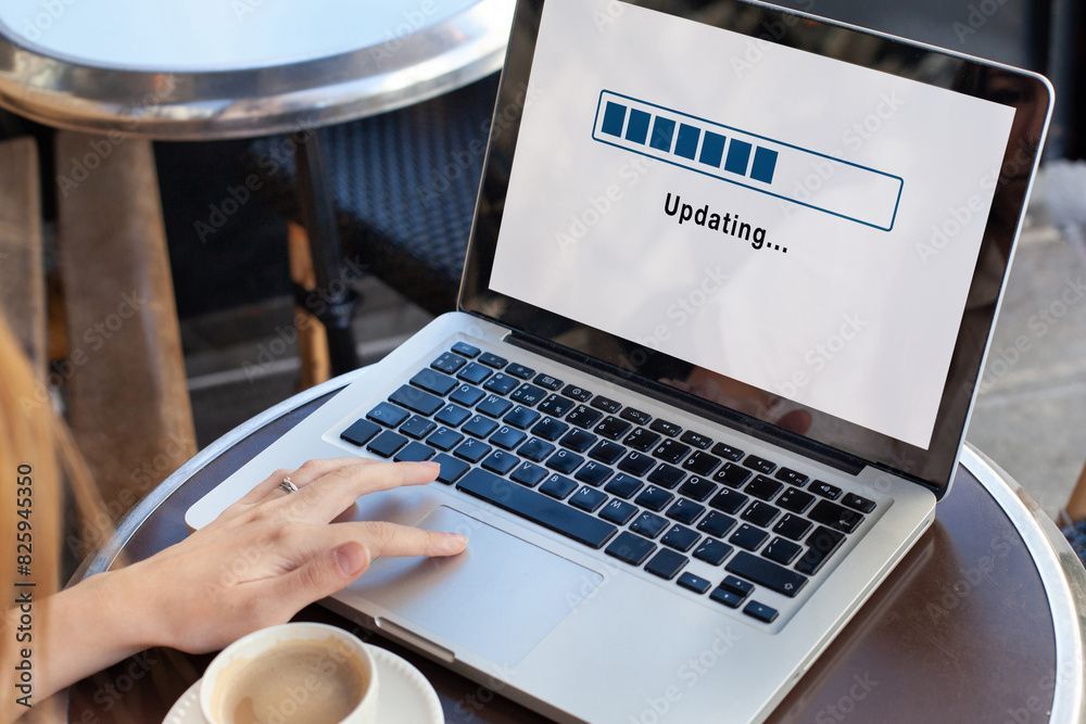 Laptop on a cafe table updating, hand on touchpad, cup of coffee, sunny outdoor setting.