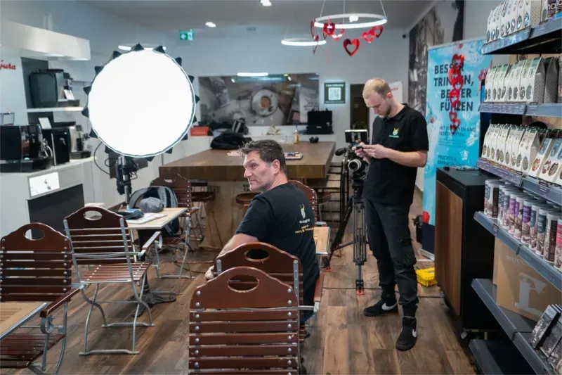 Set einer Videoproduktion: Ein Mann wird in einem Geschäft von einem Videografen mit einer Kamera gefilmt. Stativlicht im Hintergrund Bilder und verschiedene Kaffeesorten in einem Regal.