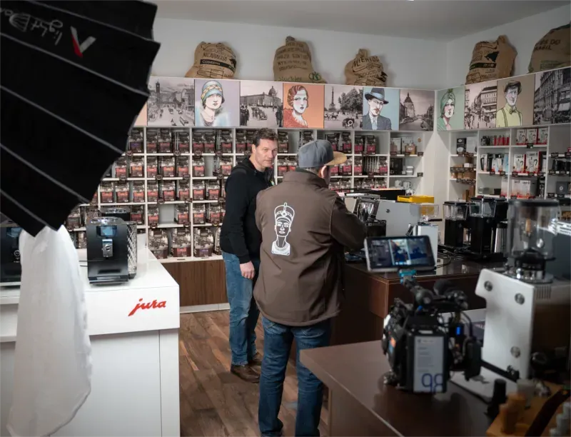 Zwei Männer in einem Kaffeemaschinengeschäft. Einer - der Videograf - filmt den anderen in der Nähe von Espressomaschinen. Kaffeebohnen in Regalen.