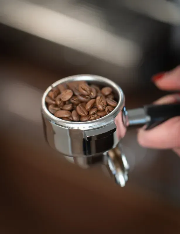 Kaffeebohnen in einem Siebträger, der von einer Person mit rot lackiertem Fingernagel gehalten wird.