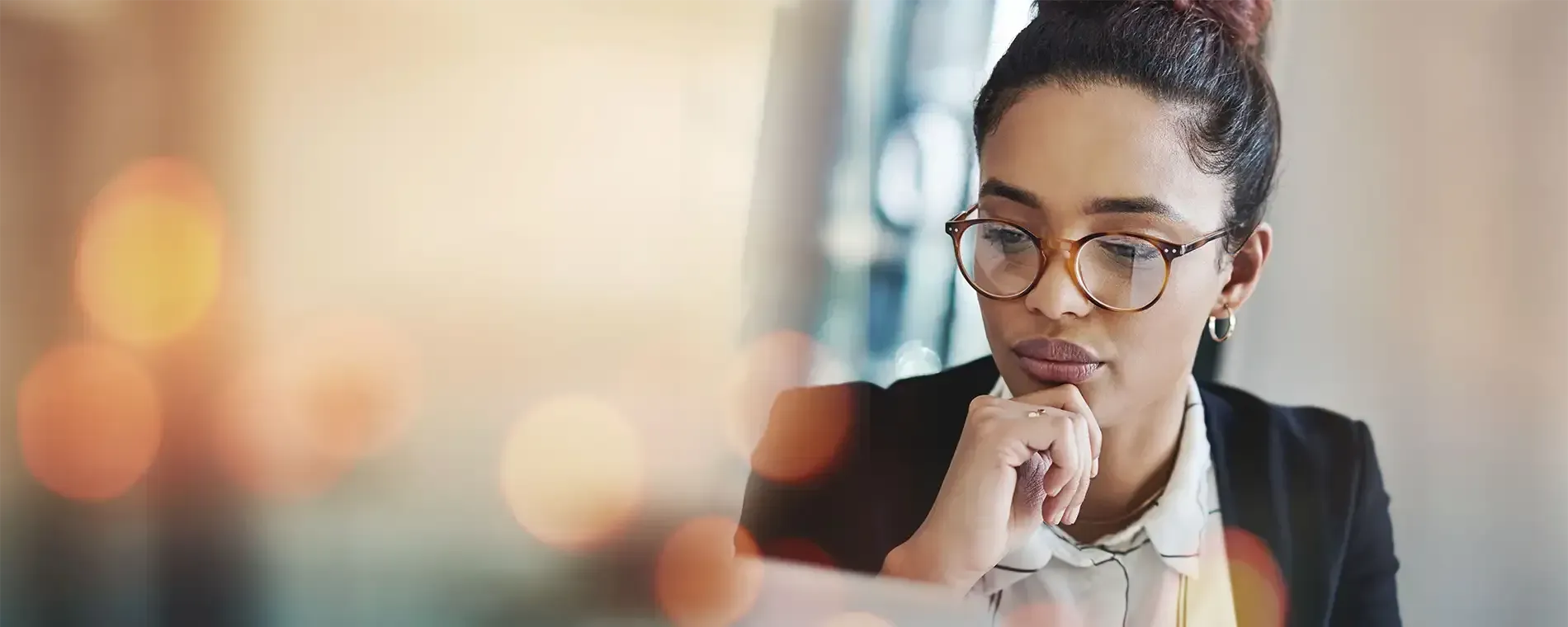 Frau mit Brille, die in einem professionellen Umfeld nachdenklich nach unten blickt, mit unscharfem Hintergrund.