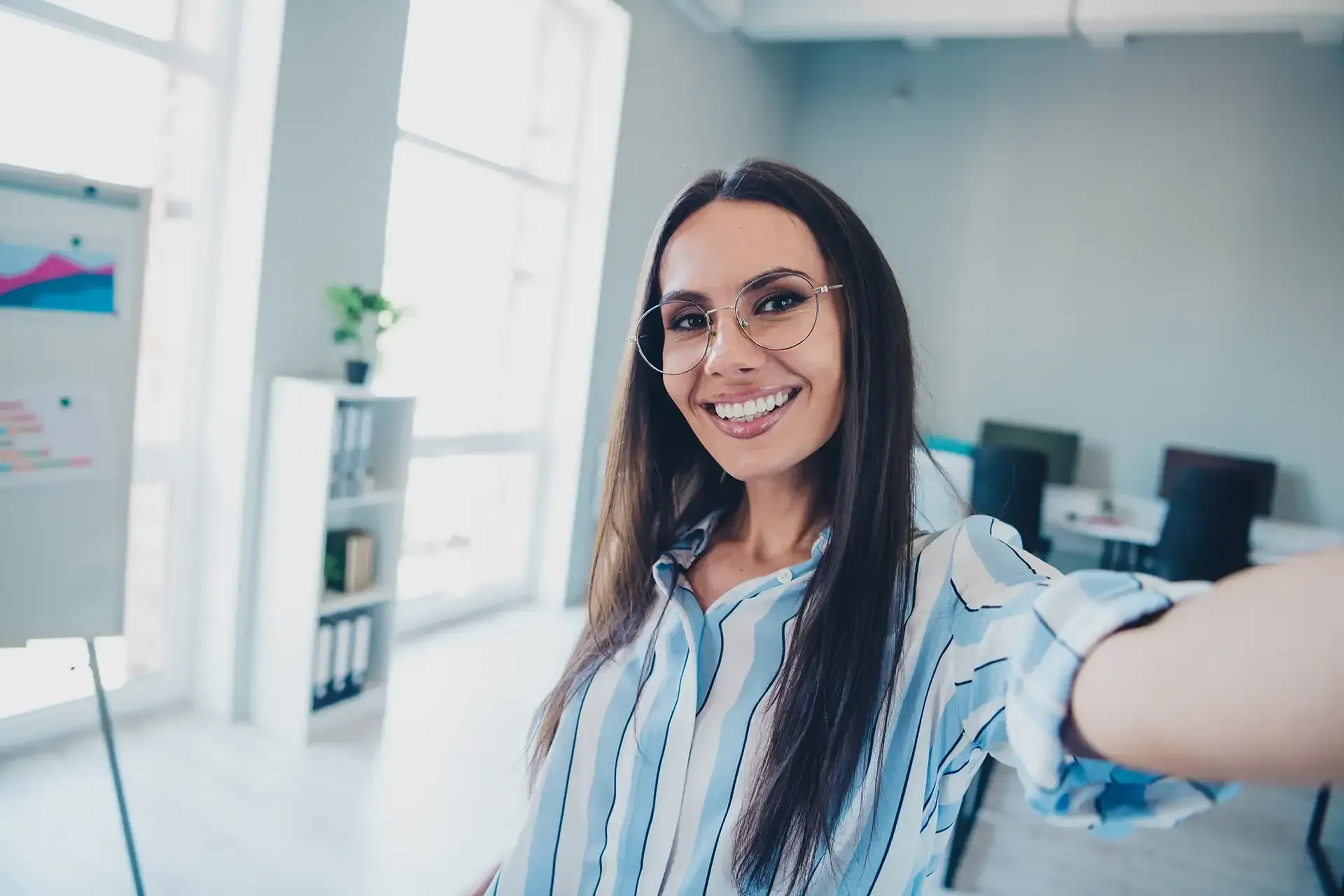 Woman smiling, taking a selfie in a bright office with a whiteboard, wearing glasses and striped shirt.