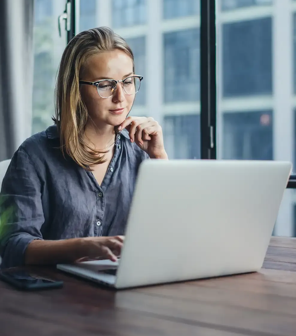 Frau mit Brille arbeitet an einem Laptop vor einem Fenster.