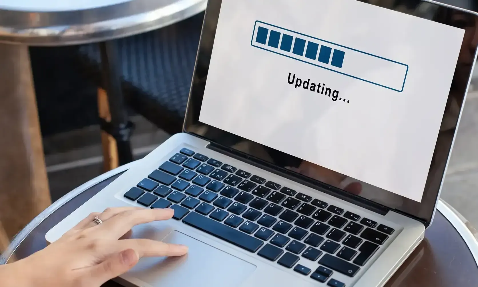 Ein Laptop zeigt auf dem Bildschirm den Hinweis „Updating…“ mit einem Fortschrittsbalken. Er steht auf einem kleinen Tisch, während eine Hand gerade auf der Tastatur tippt.