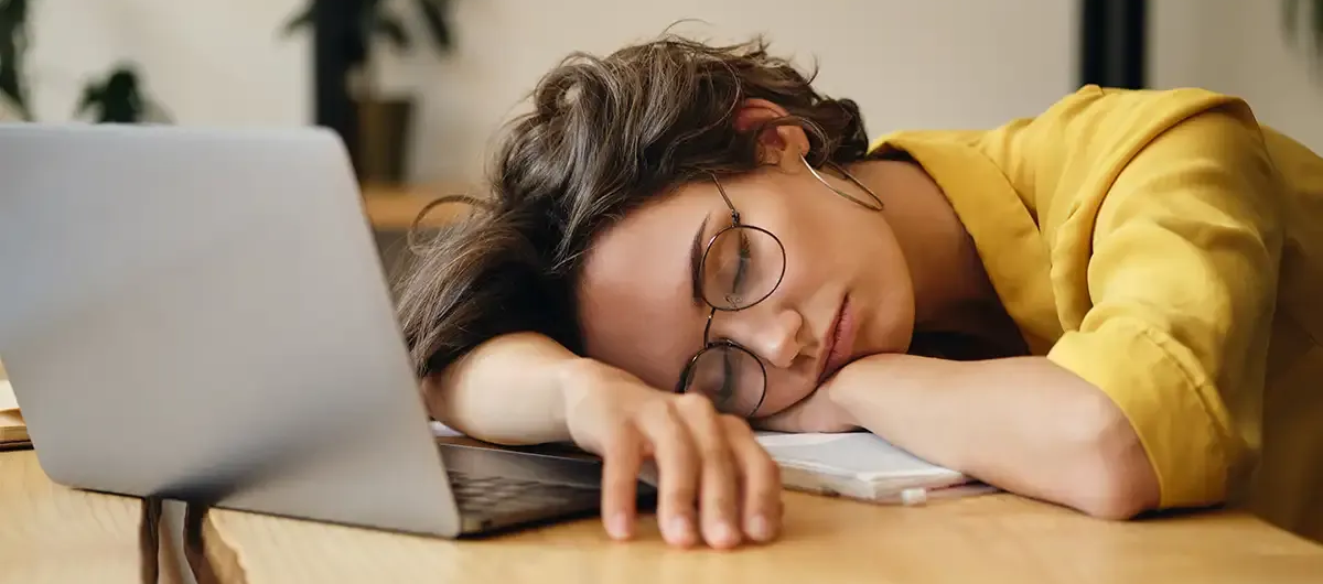 Eine Frau schläft an ihrem Schreibtisch, den Kopf auf die Arme neben einem Laptop gelegt. Sie trägt eine Brille und ein gelbes Hemd.