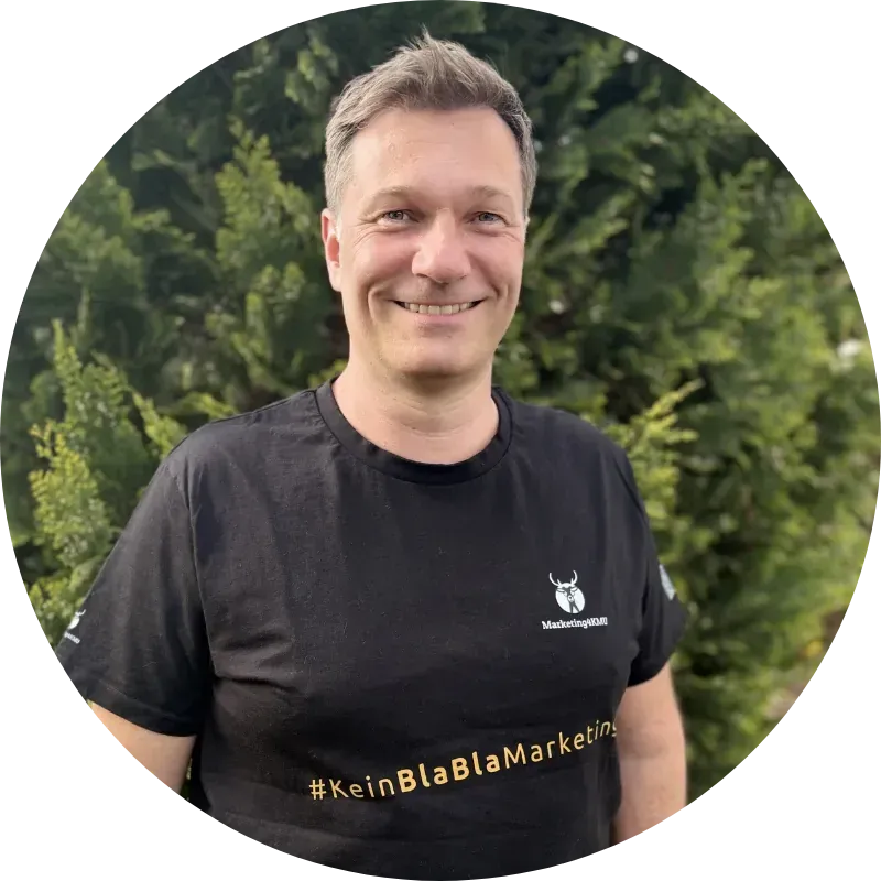 Kay Albusberger | Marketing4KMU Eine lächelnde Person in einem schwarzen T-Shirt steht im Freien vor grünen Bäumen.