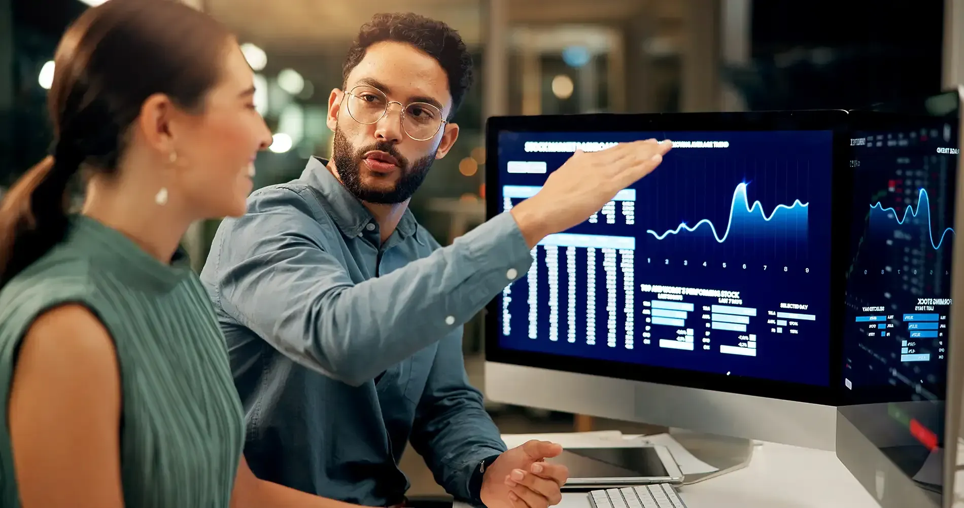 Ein Mann zeigt auf Daten auf einem Monitor und bespricht diese mit einer Frau in einer Büroumgebung.