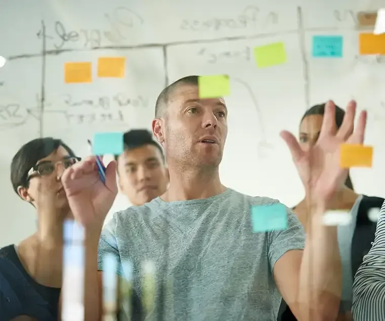 Gruppe von Menschen beim Brainstorming, die Haftnotizen an einer Glaswand betrachten. Ein Mann gestikuliert, die anderen schauen zu.