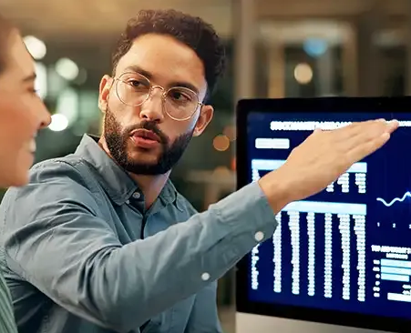 Ein Mann zeigt auf einen Computerbildschirm und erklärt einer Person Daten. Sie befinden sich in einem Büro.