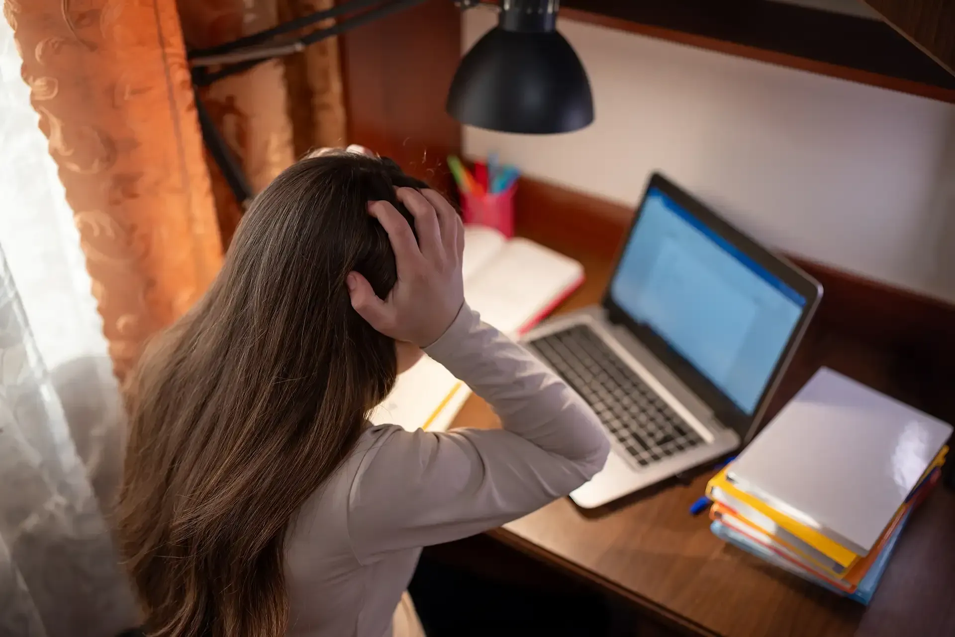 Girl with long brown hair, hands on her head, looks stressed, sits at a desk with a laptop and papers.