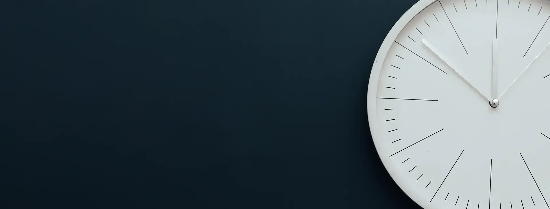 White clock on a dark blue wall. The clock hands show it is close to 10:00.