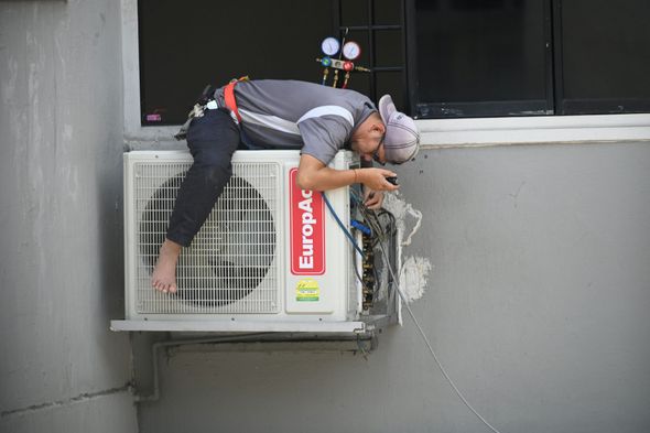 Person lying on an outdoor air conditioning unit, working with gauges, gray building background.