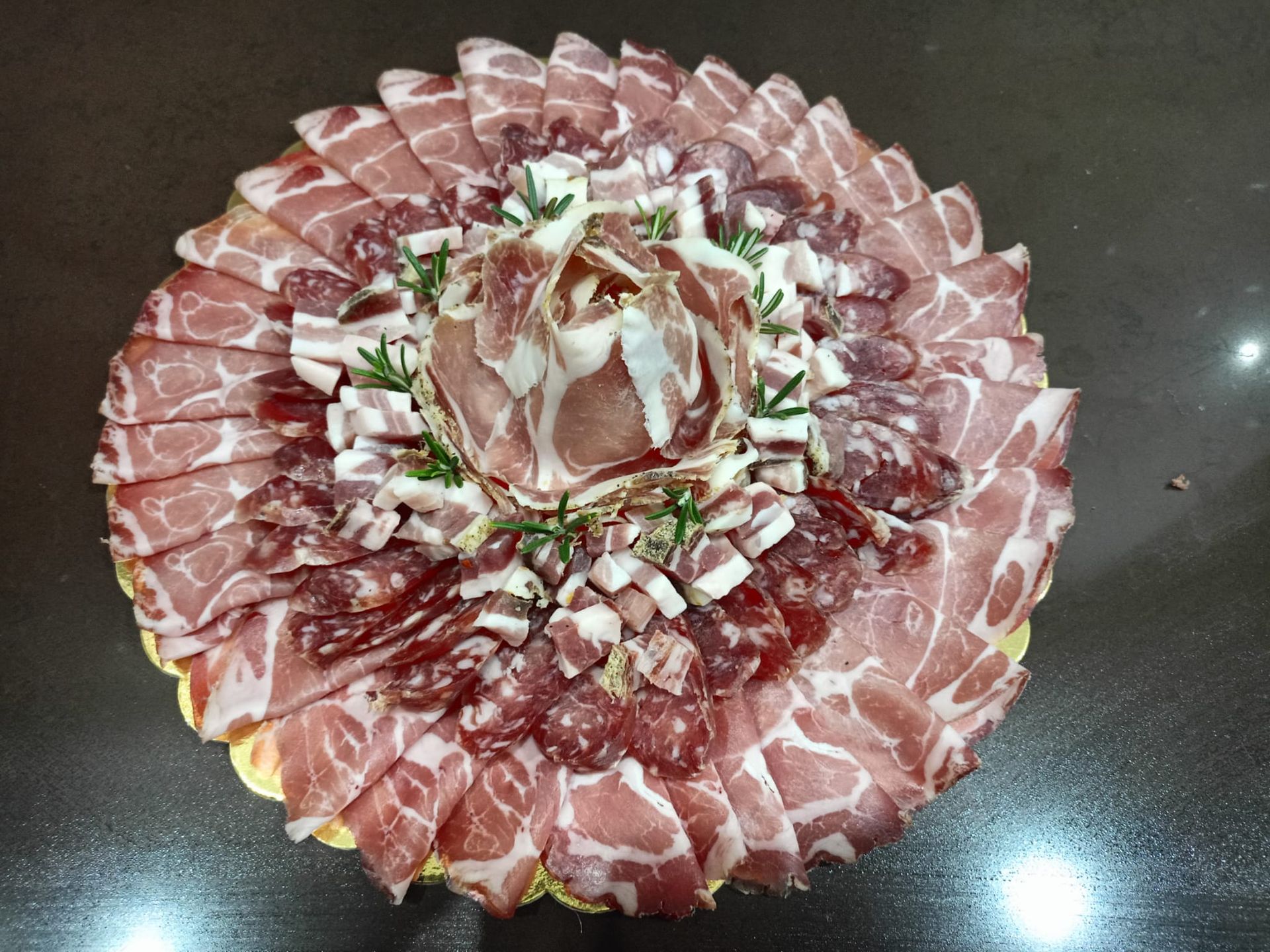 Piatto a forma di fiore con affettati sottili e guarnito con erbe aromatiche, su un tavolo scuro.