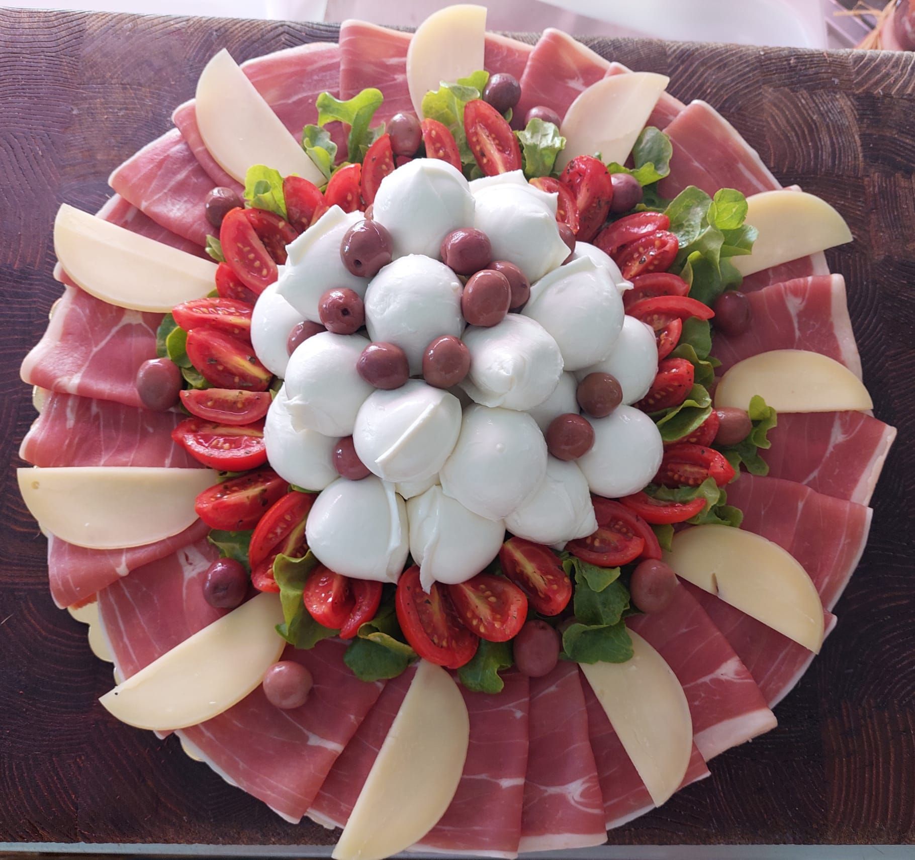 Piatto misto di salumi con prosciutto crudo, fragole, mozzarella, uva e fette di pera disposte a formare un motivo floreale.