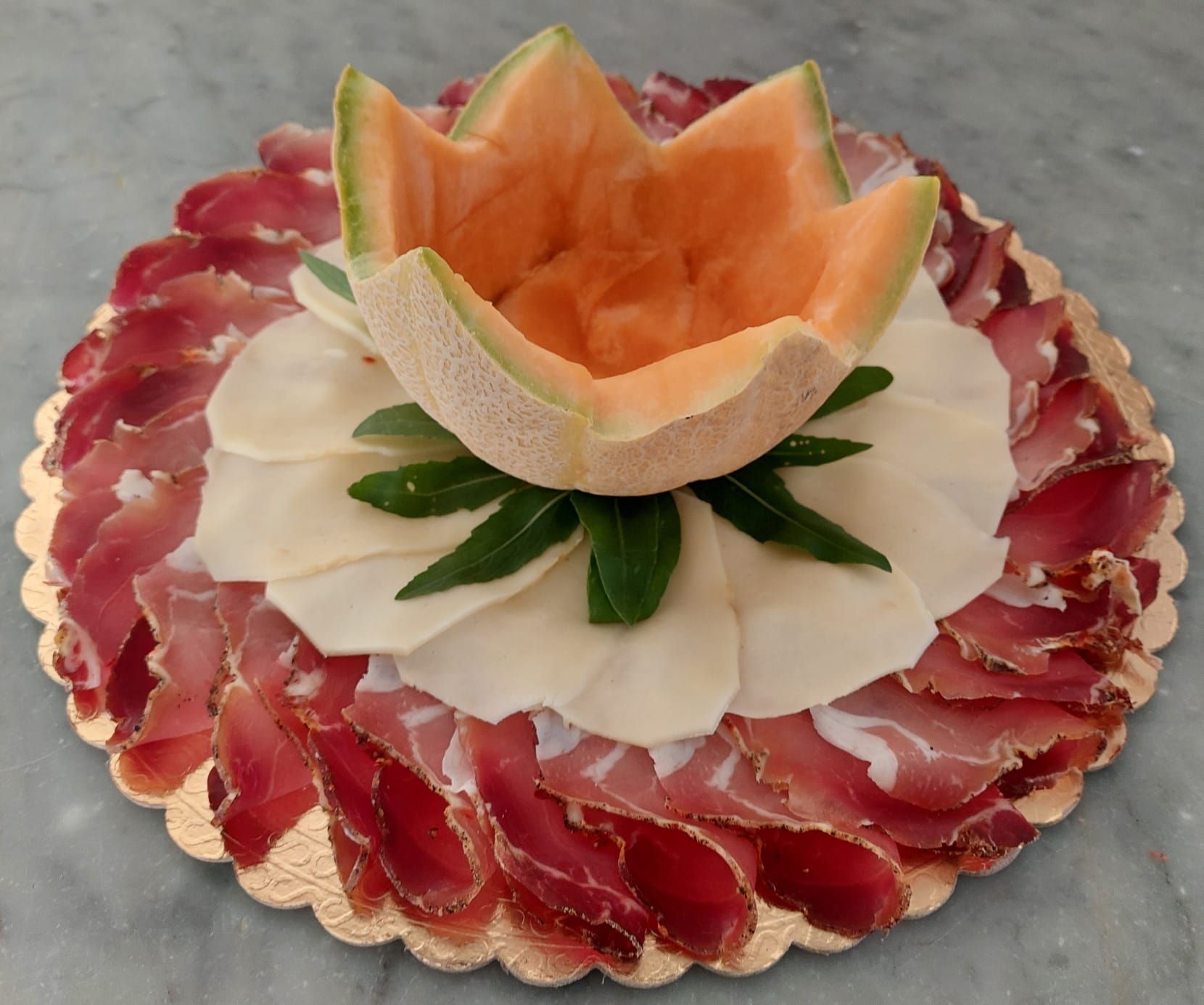 Piatto gourmet con salumi tagliati finemente e disposti ad anello attorno a fiori di melone e formaggio bianco.