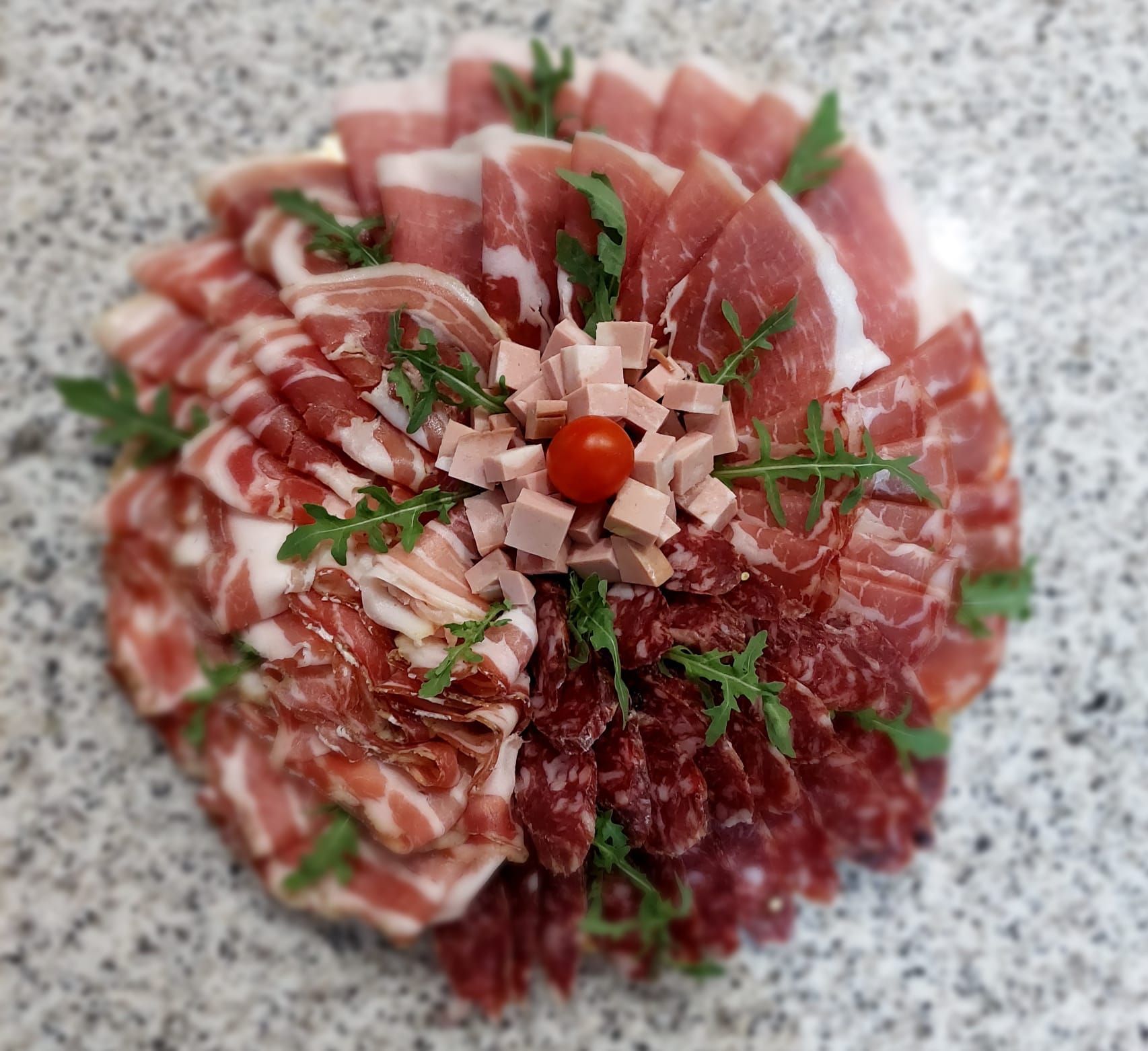 Affettati assortiti disposti a forma di fiore su un piatto con prezzemolo e un pomodorino ciliegino al centro.