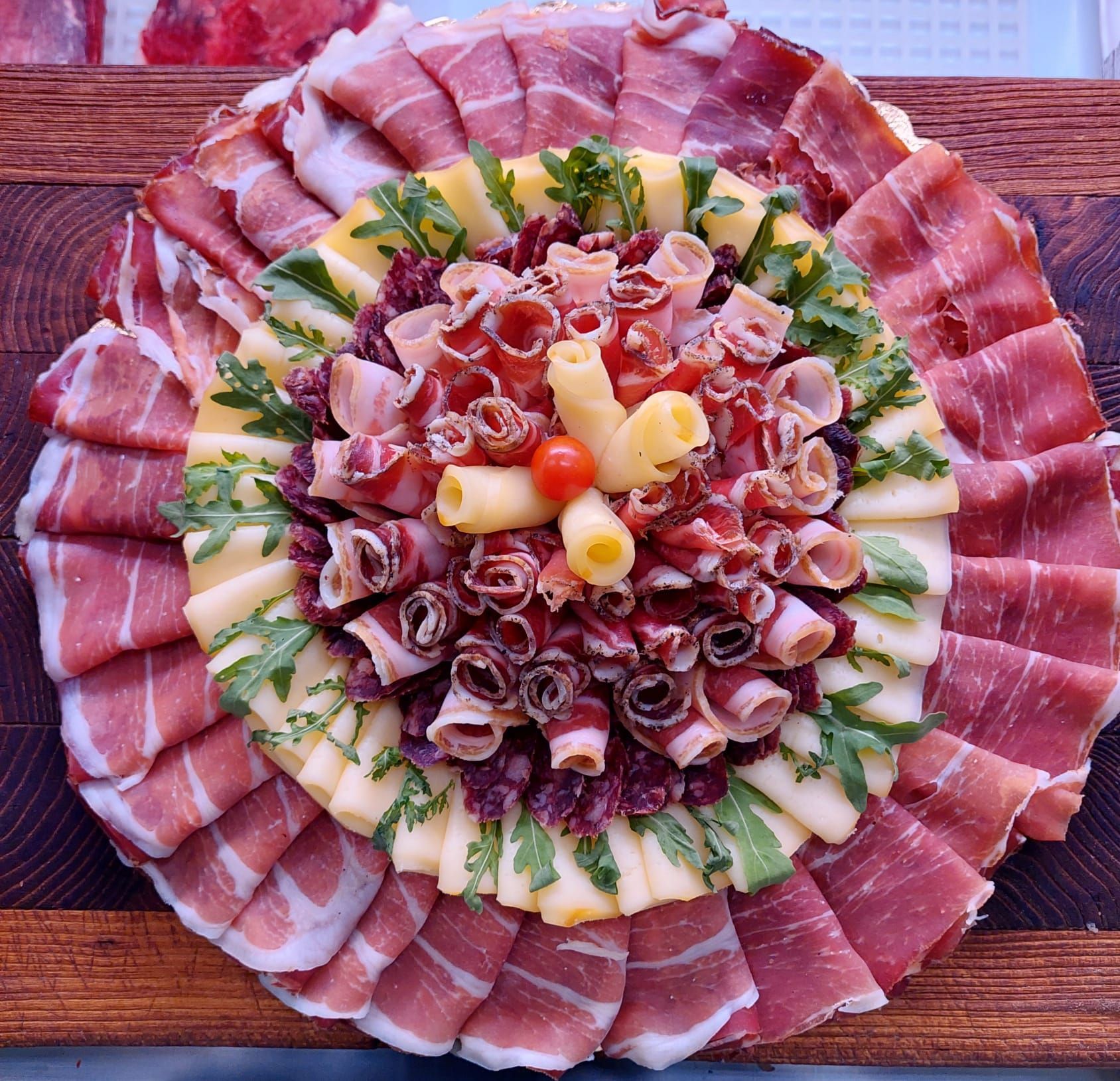 Affettati assortiti disposti in anelli concentrici su un tagliere di legno, con guarnizione di formaggio ed erbe aromatiche al centro.