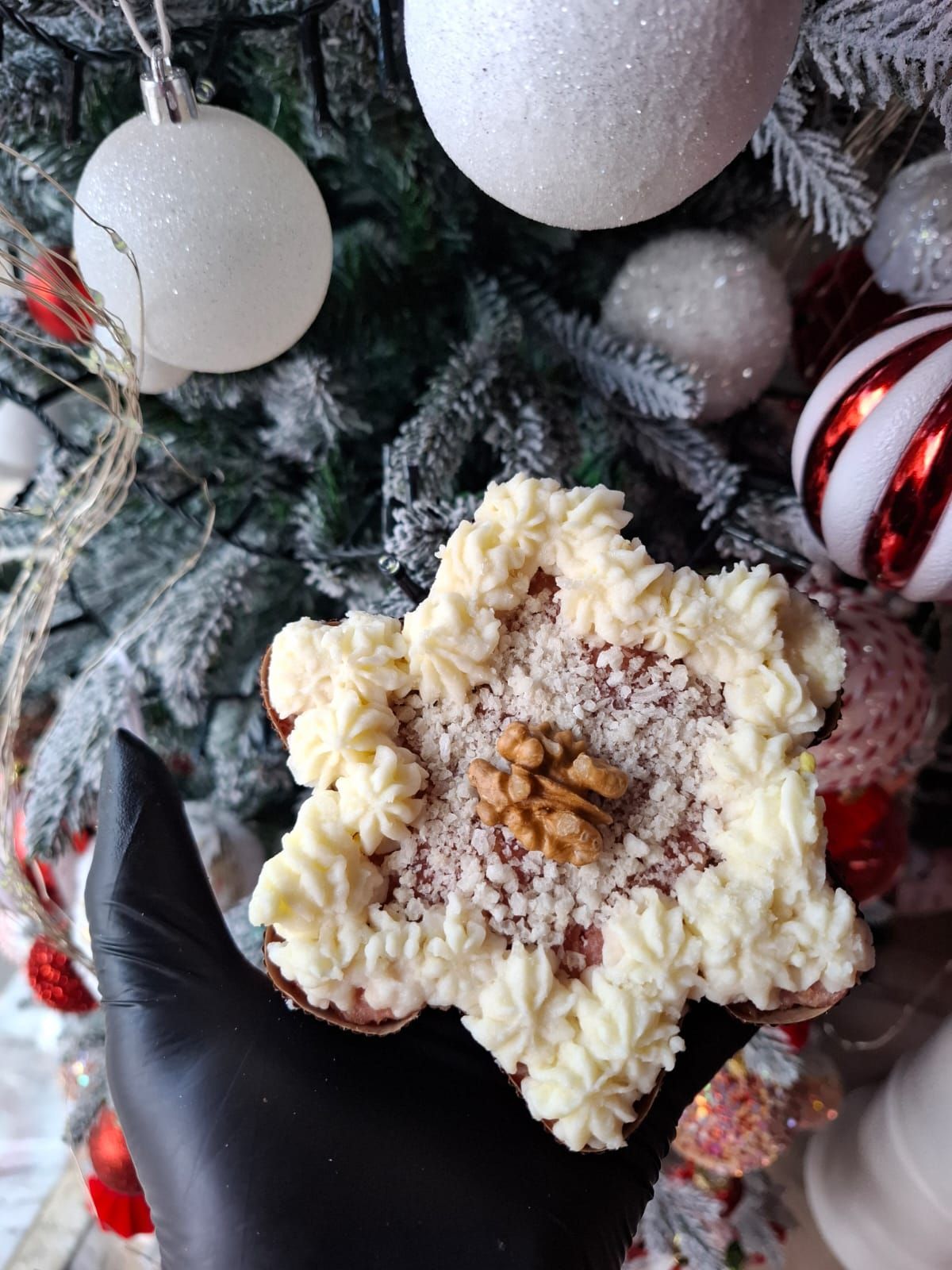 Una mano guantata di nero regge un biscotto a forma di fiocco di neve con glassa alla crema e zuccherini colorati accanto a un albero di Natale decorato.