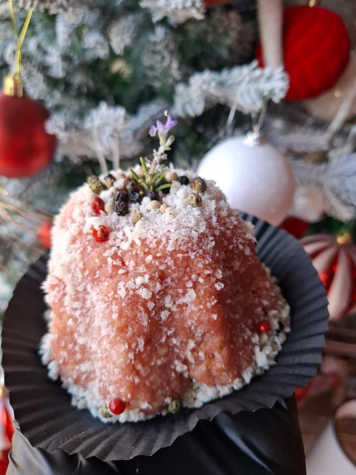 Dessert rosa ricoperto di zucchero, avvolto in un involucro nero e decorato con una guarnizione verde, posto davanti a un albero di Natale.