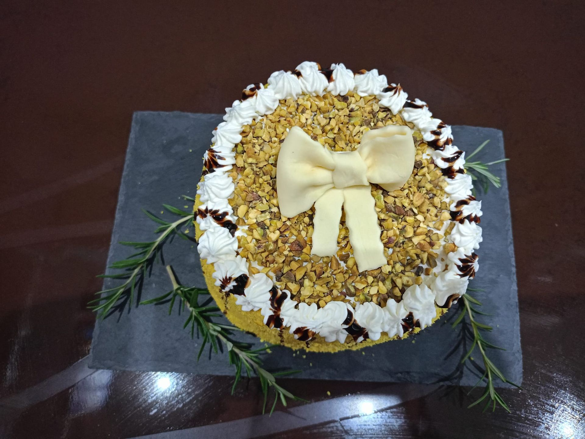 Dessert rotondo decorativo sormontato da un fiocco di crema e fiori su un piatto di ardesia nera con verde.