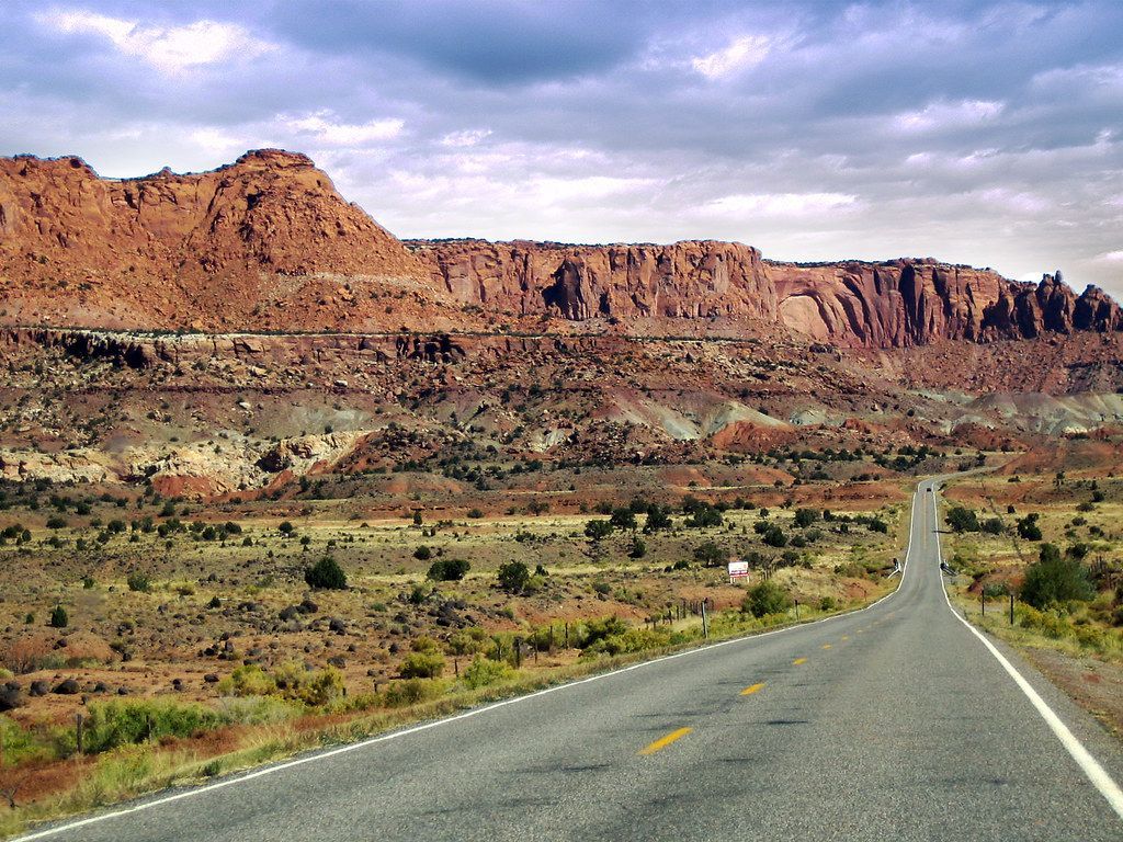 A straight road leads toward distant red rock cliffs and mesas under a cloudy blue sky in a desert landscape.