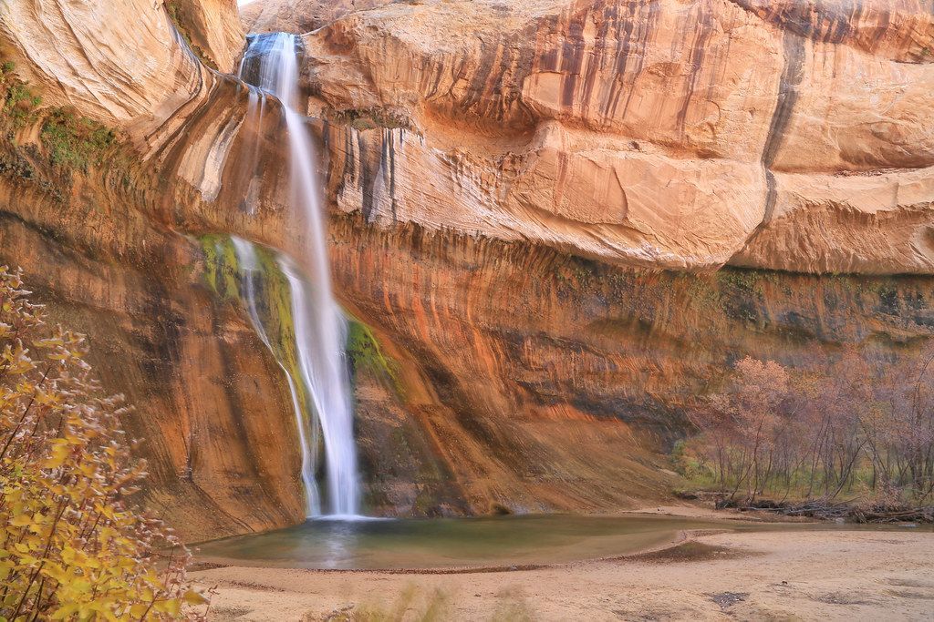 A slender waterfall cascades down a massive, curved sandstone cliff into a small, calm pool in a desert canyon.