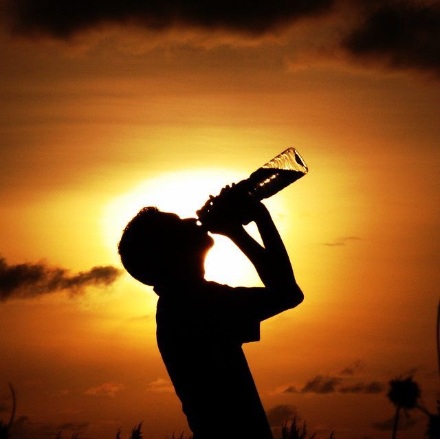 Silhouette of a person drinking from a bottle against a bright, warm orange sunset.