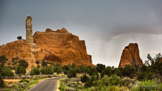 A paved road leads toward tall, weathered orange rock formations under a cloudy, overcast sky.