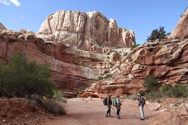 Three hikers with backpacks stand on a red dirt trail beneath towering, layered sandstone cliffs under a clear blue sky.