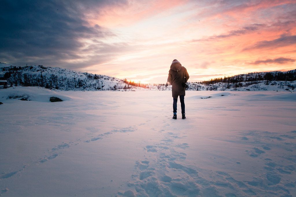 A person standing alone on a vast, snow-covered landscape under a colorful sunrise sky with footprints leading away.