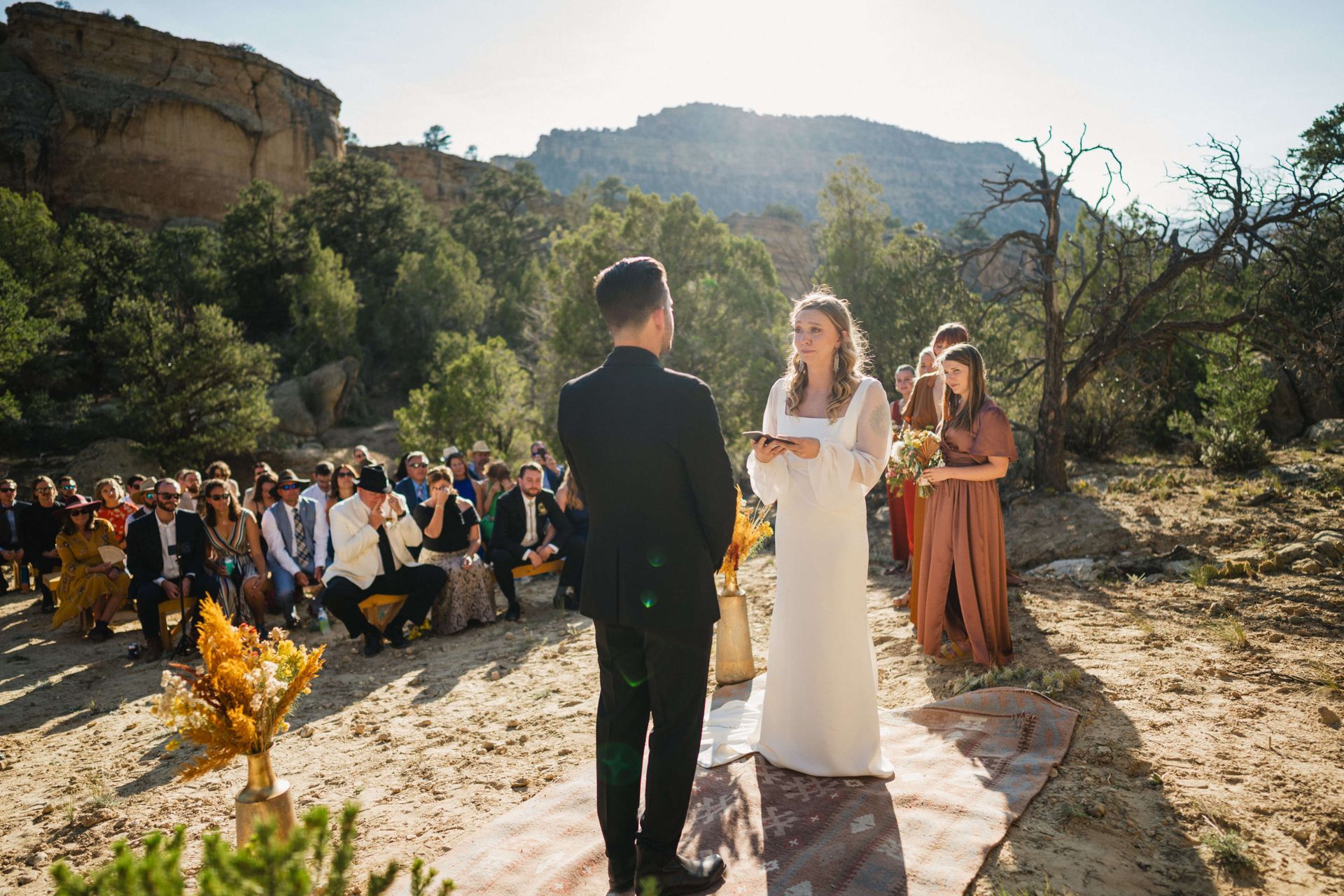 A couple stands facing each other during an outdoor wedding ceremony in a desert landscape with guests seated behind.