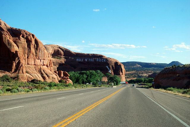 A road stretches toward a massive red sandstone cliff in the desert under a clear blue sky.