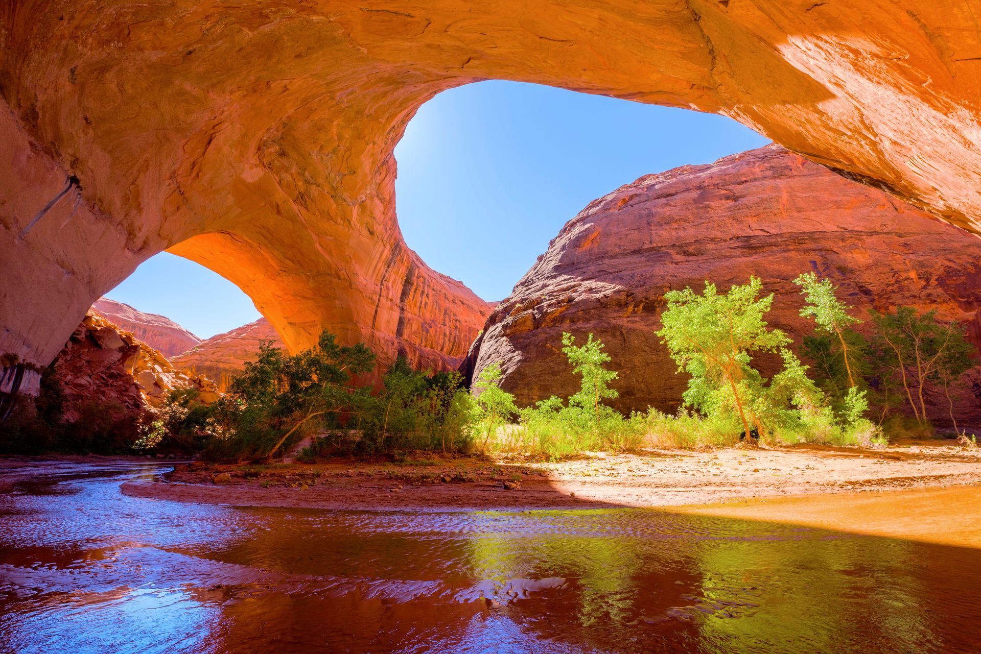 A stream flows beneath the towering, curved rock arch of Coyote Gulch, framed by bright green trees and red canyon walls.