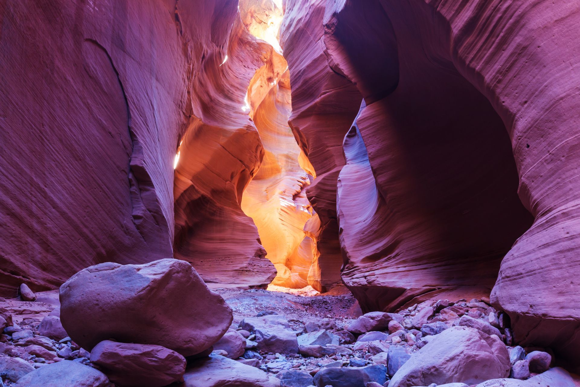 A narrow slot canyon with smooth, wavy walls in shades of purple and orange, illuminated by light at the far end.
