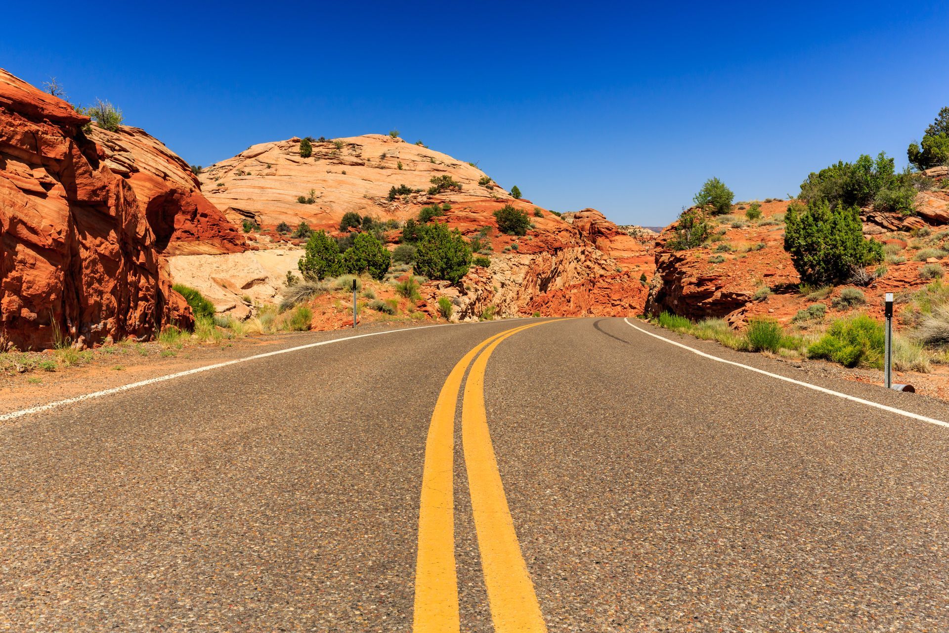 A paved road with yellow center lines cuts through a desert landscape of red rock formations under a bright blue sky.