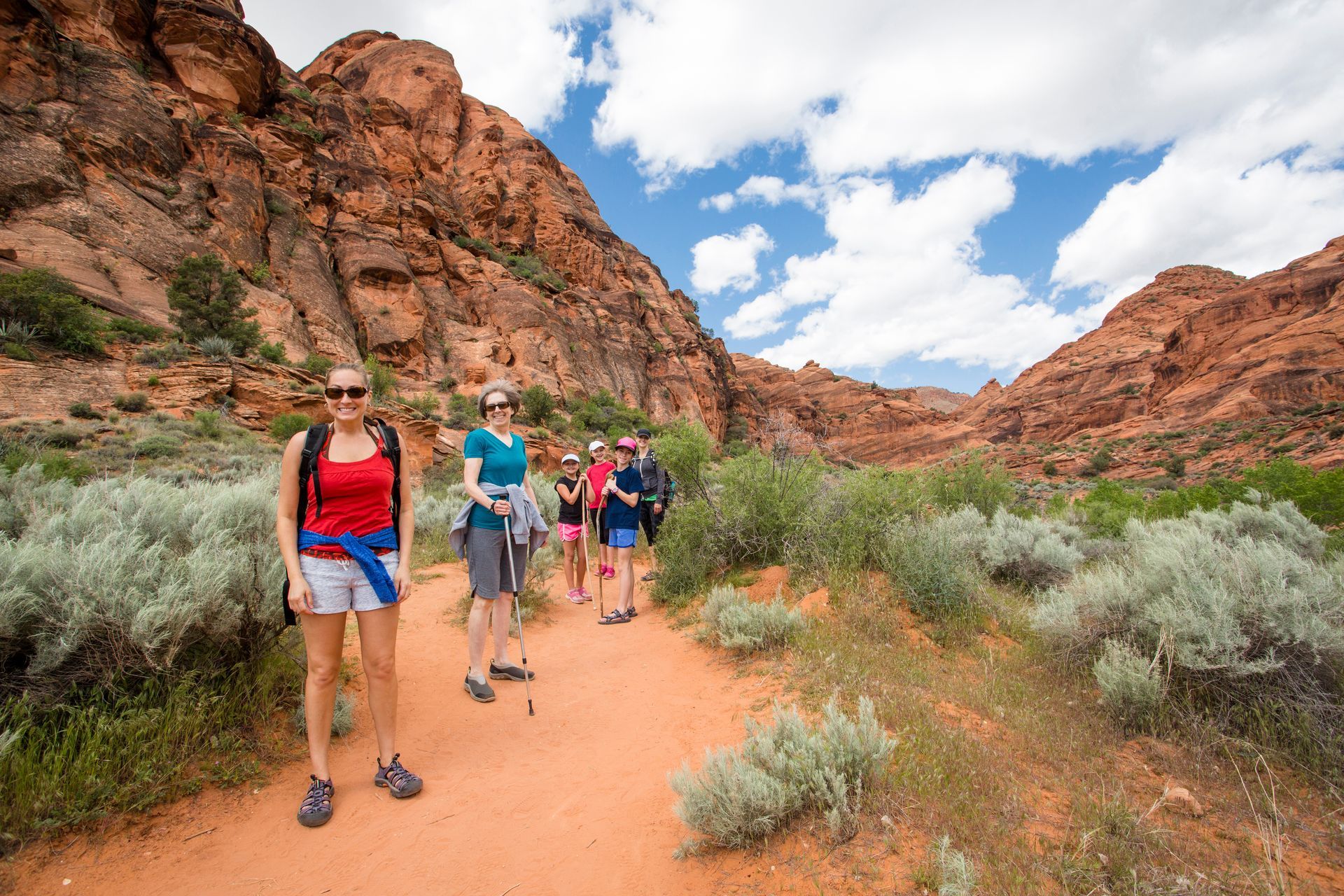 A group of hikers stands on a red dirt trail surrounded by desert shrubs and towering red rock cliffs under a cloudy sky.