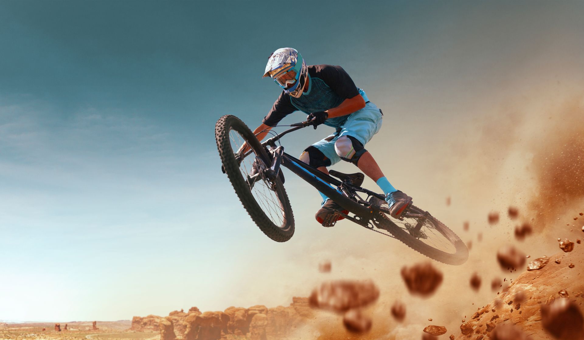 A mountain biker in protective gear performs an airborne stunt over a dusty, rocky terrain under a bright blue sky.