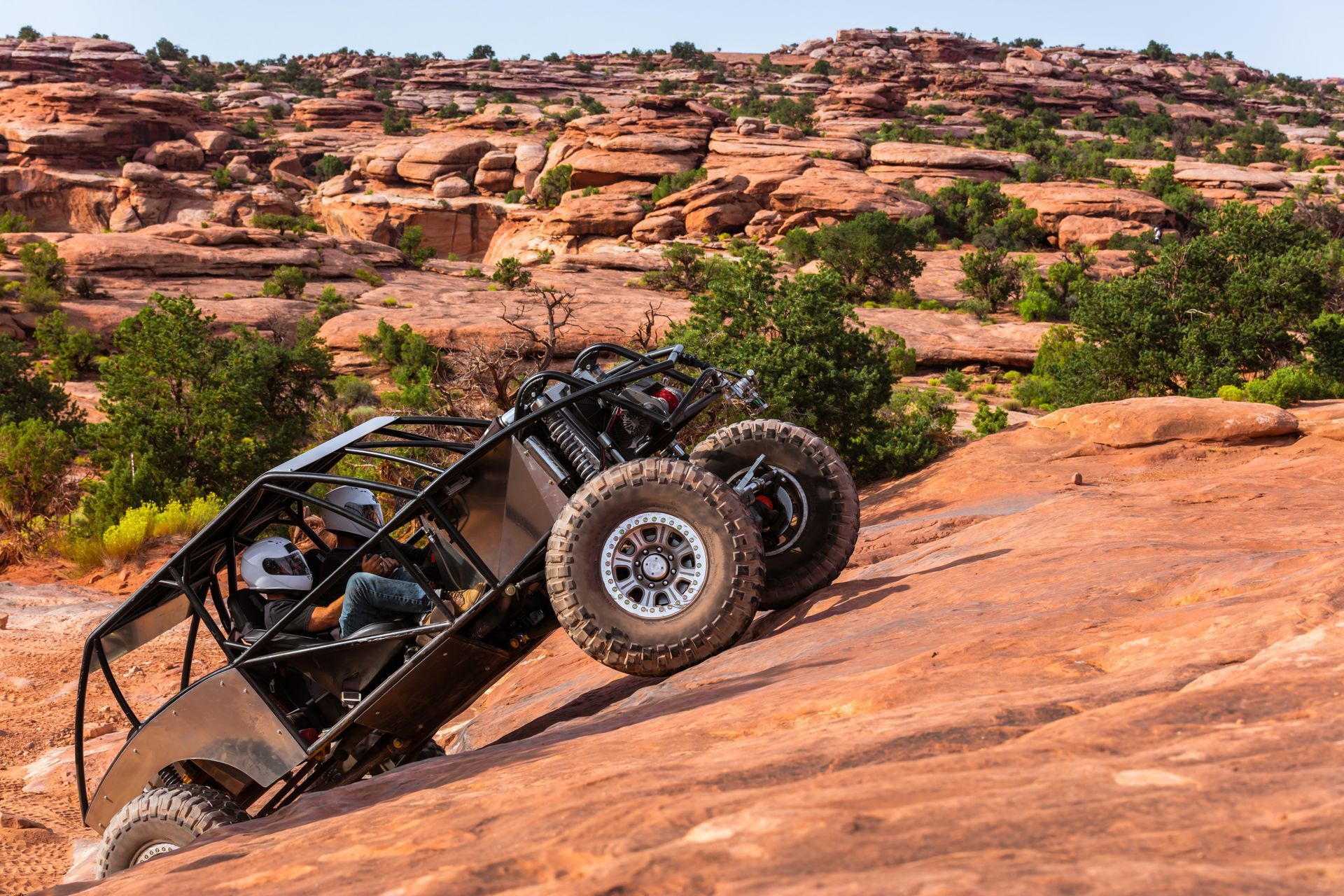 A black off-road vehicle climbs a steep red rock slope in a desert landscape.