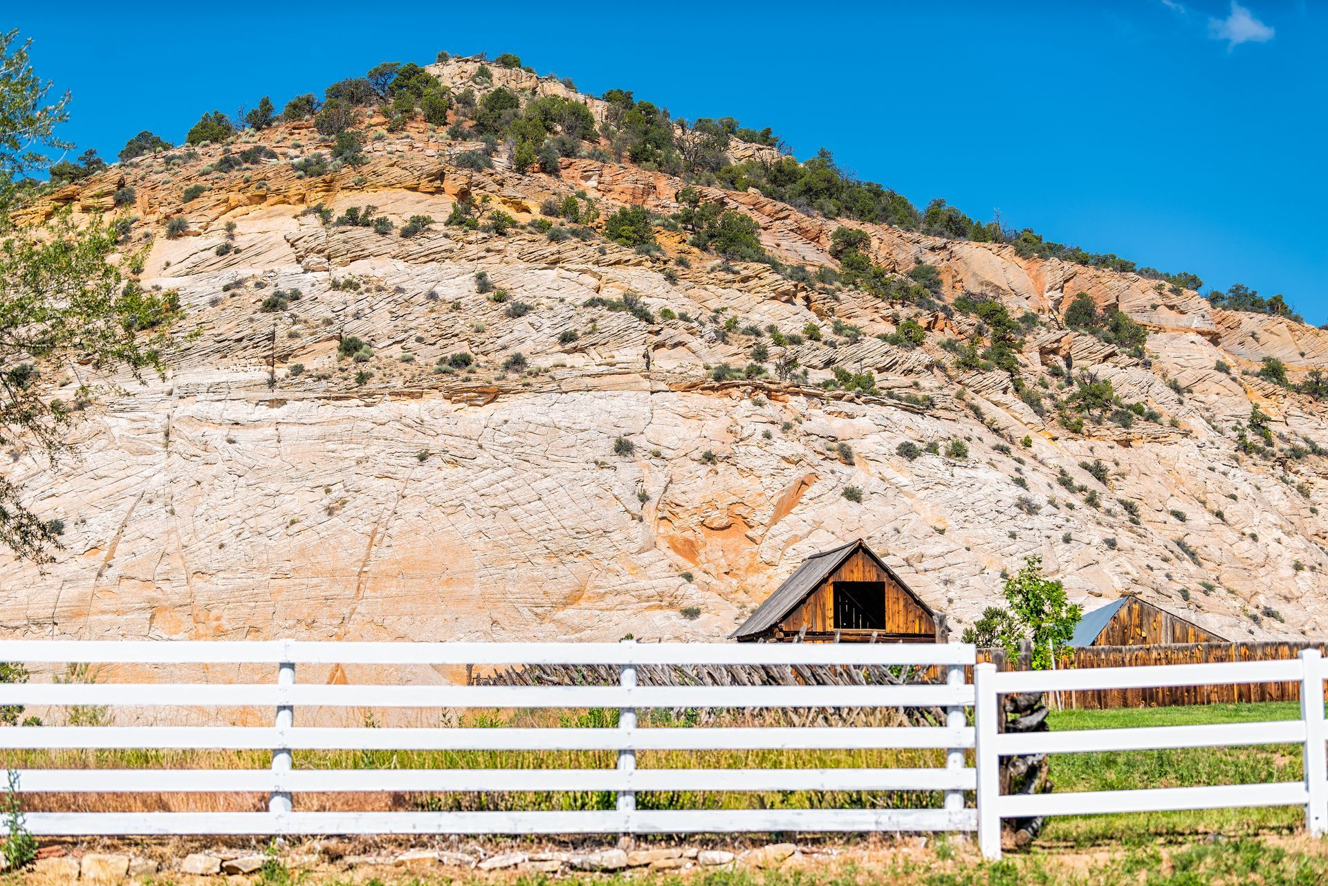 A white fence sits before a wooden barn against a large, rugged, tan-colored mountain under a clear blue sky.